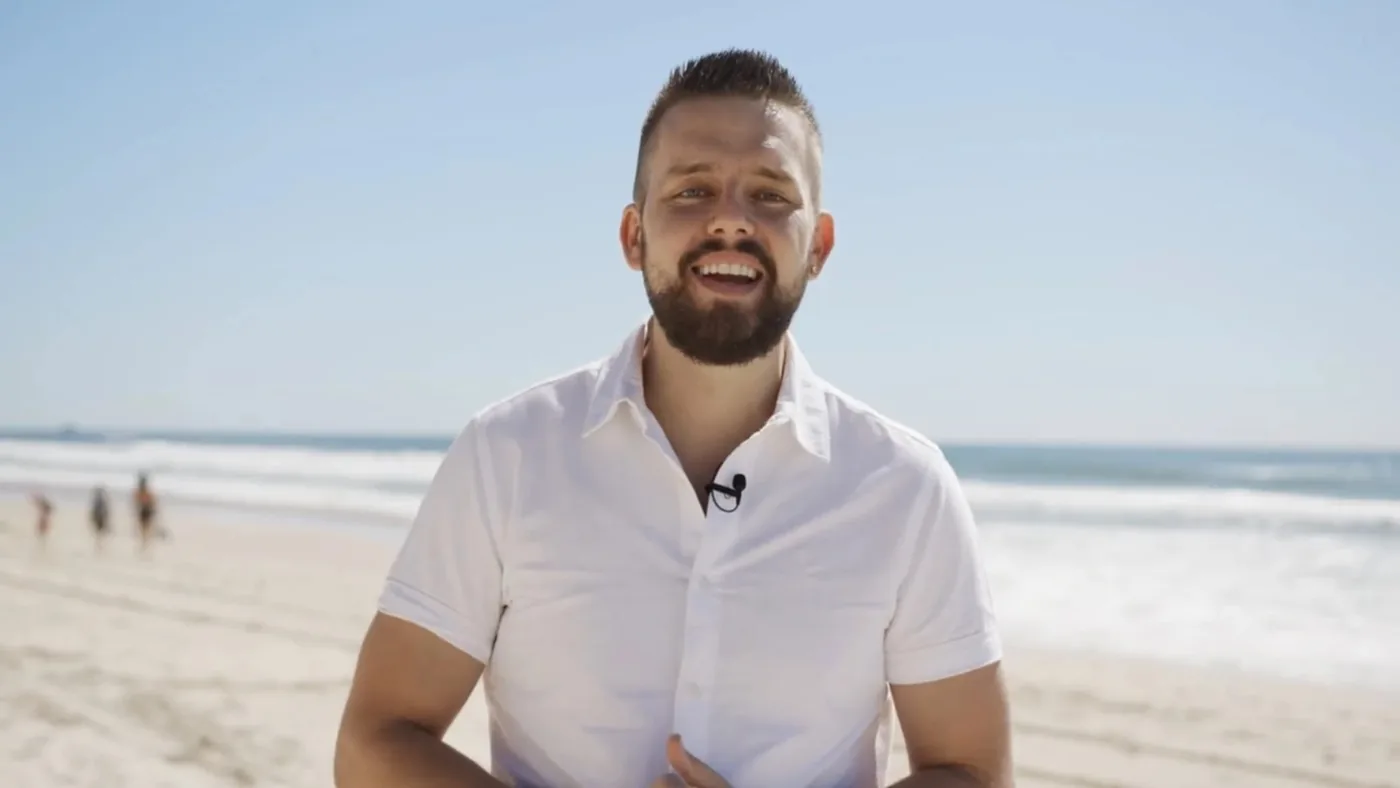 Luke on the beach in a white shirt, talking to camera